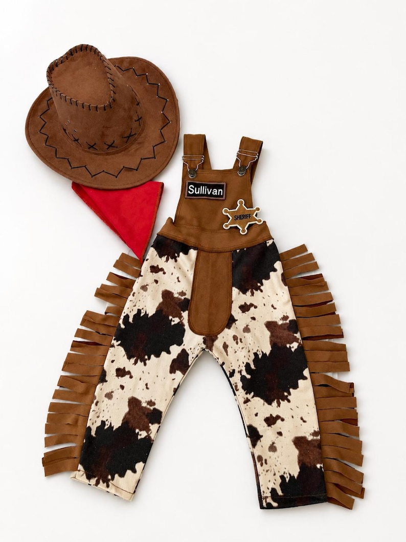 Op de afbeelding: Een bruine cowboyhoed, een rode bandana en een jumpsuit met koeienprint, franjes en een sheriff-badge. Op de jumpsuit staat de naam "Sullivan". Deze western-outfit is perfect voor een kostuum.