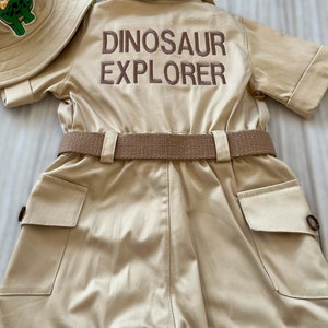 Pu&ograve; includere: Una tuta di stile safari color beige con maniche corte e una cintura marrone. La tuta ha due grandi tasche sul davanti e le parole "Dinosaur Explorer" ricamate sul retro con filo marrone.