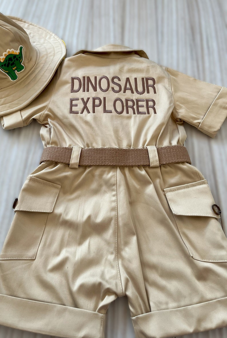 Pu&ograve; includere: Una tuta di stile safari color beige con maniche corte e una cintura marrone. La tuta ha due grandi tasche sul davanti e le parole "Dinosaur Explorer" ricamate sul retro con filo marrone.