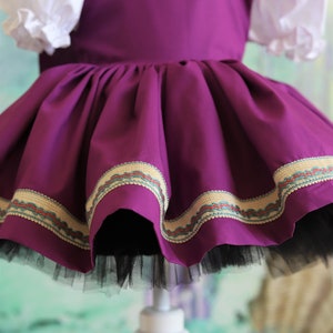 Peut inclure: Une robe violette avec un corsage blanc et une jupe en tulle noire. La robe est orn&eacute;e d'une bordure dor&eacute;e et verte autour du bas.