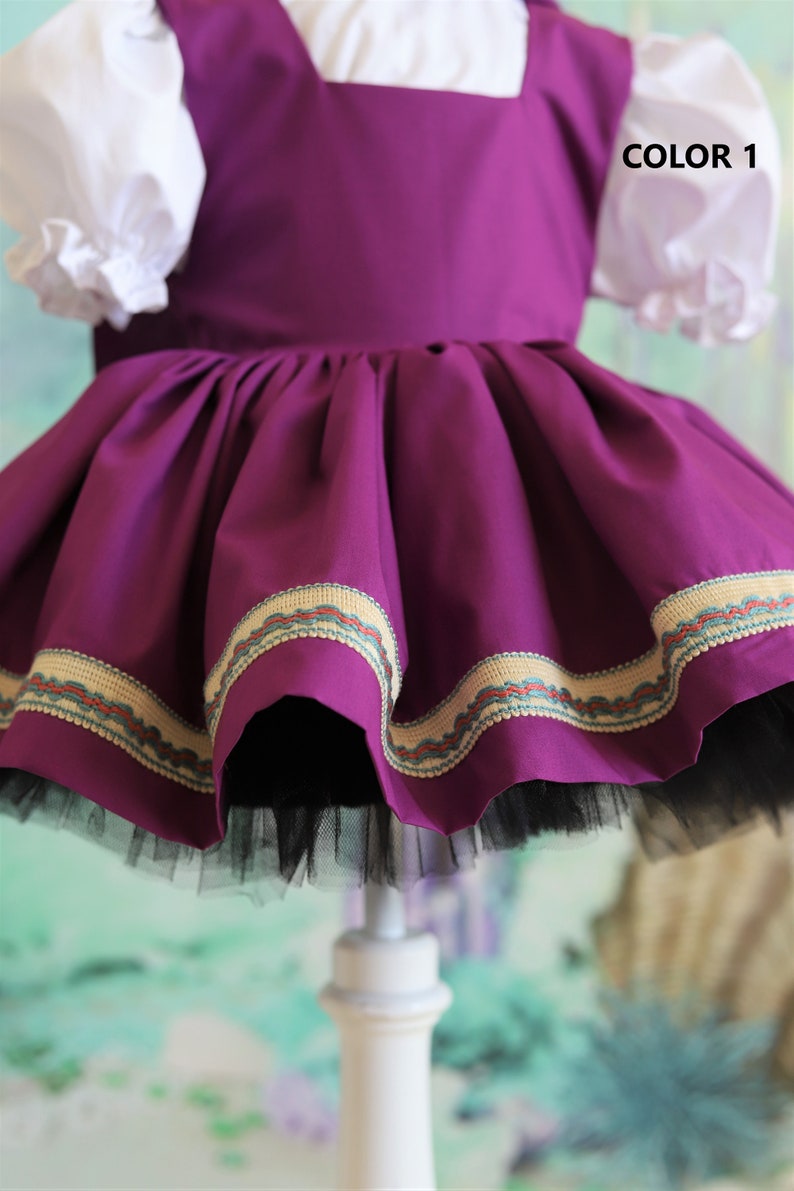 Peut inclure: Une robe violette avec un corsage blanc et une jupe en tulle noire. La robe est orn&eacute;e d'une bordure dor&eacute;e et verte autour du bas.