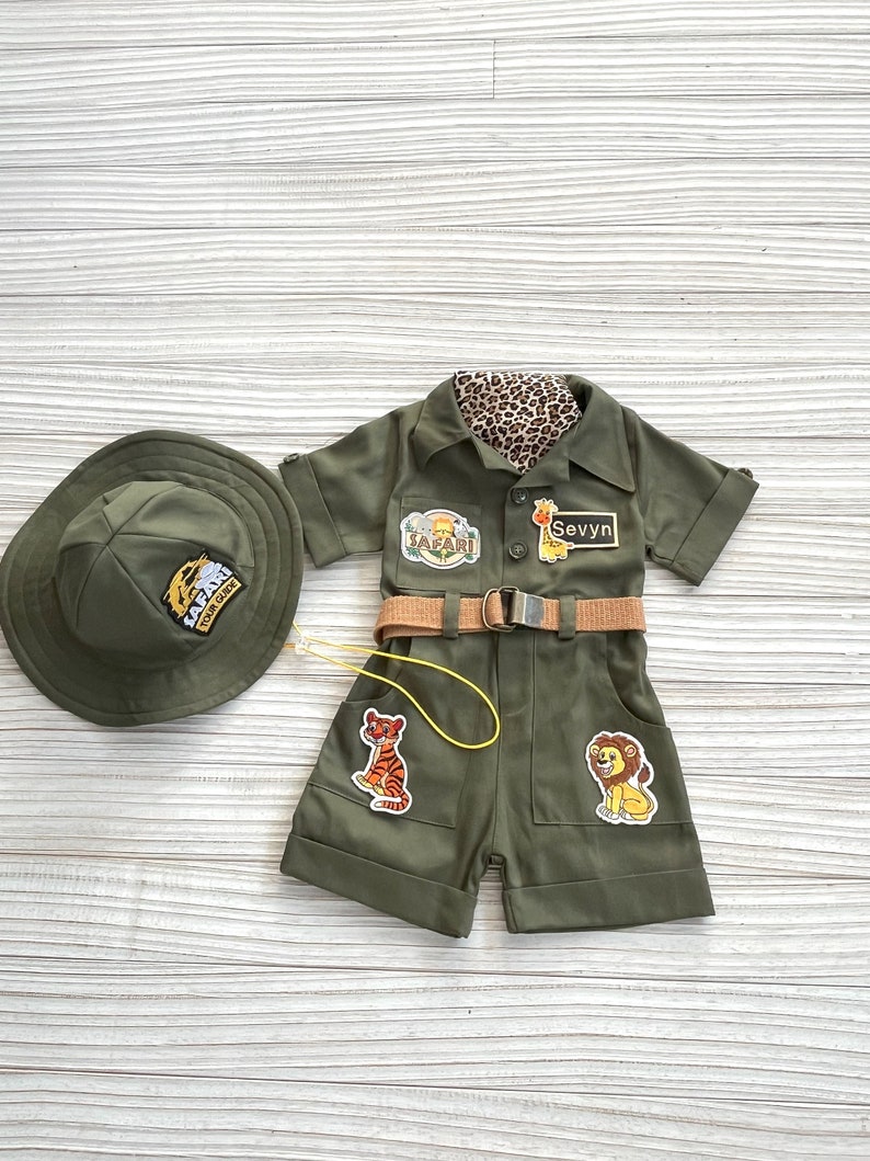 May include: A green safari-themed outfit for a child, featuring a hat, a short-sleeved jumpsuit with pockets, a belt, and a yellow lanyard. The jumpsuit has patches with animal illustrations and the text "Safari" and "Sevyyn".