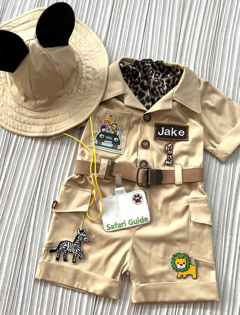 May include: A tan safari outfit for a child with a safari guide badge, a zebra patch, a lion patch, and a jeep patch. The outfit includes a hat with Mickey Mouse ears, a short-sleeved shirt, and shorts with a belt. The name "Jake" is written on a name tag.