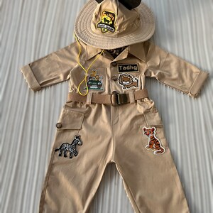 May include: A tan safari jumpsuit with a brown belt and multiple patches, including a zebra, a tiger, a lion, and a jeep. The jumpsuit has a yellow cord attached to the collar. The outfit also includes a tan safari hat with Mickey Mouse ears and a patch that says "Safari Guide". The name "Tadhg" is written on a patch on the chest.