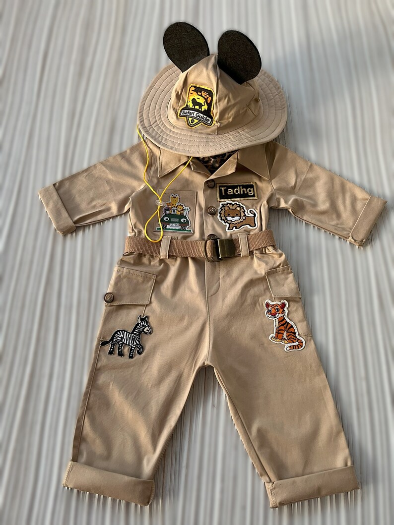 May include: A tan safari jumpsuit with a brown belt and multiple patches, including a zebra, a tiger, a lion, and a jeep. The jumpsuit has a yellow cord attached to the collar. The outfit also includes a tan safari hat with Mickey Mouse ears and a patch that says "Safari Guide". The name "Tadhg" is written on a patch on the chest.