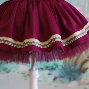 Peut inclure: Une jupe en tulle bordeaux avec une bordure d&eacute;corative autour de l'ourlet. La bordure est une combinaison de beige, marron et bleu.