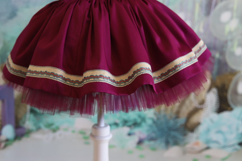 Peut inclure: Une jupe en tulle bordeaux avec une bordure d&eacute;corative autour de l'ourlet. La bordure est une combinaison de beige, marron et bleu.