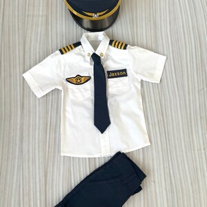 May include: A child's pilot costume set, including a navy blue hat with gold trim, a white short-sleeved shirt with gold epaulets and a navy tie, and navy blue pants. The shirt has a patch that says "Jaxson".
