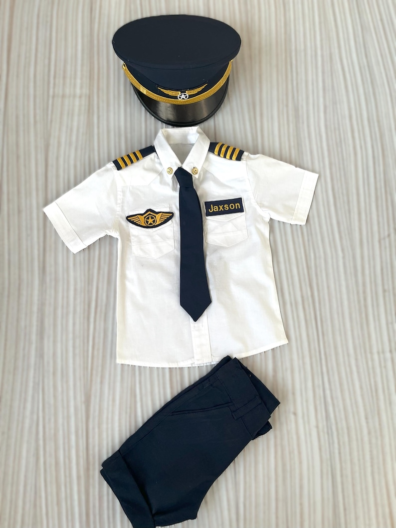 May include: A child's pilot costume set, including a navy blue hat with gold trim, a white short-sleeved shirt with gold epaulets and a navy tie, and navy blue pants. The shirt has a patch that says "Jaxson".
