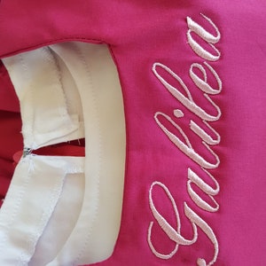 K&ouml;nnte beinhalten: Ein rosa Kleid mit einem wei&szlig;en Kragen und dem Wort "Gabriela" in rosa Stickerei auf der Vorderseite.