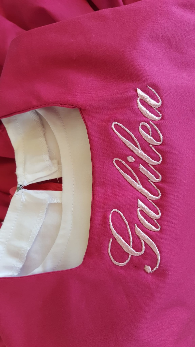 K&ouml;nnte beinhalten: Ein rosa Kleid mit einem wei&szlig;en Kragen und dem Wort "Gabriela" in rosa Stickerei auf der Vorderseite.