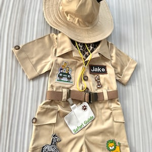 May include: A tan safari outfit for a child with a safari hat, a name tag that says "Jake", and a safari guide badge. The outfit has patches of a zebra, a lion, and a jeep with animals on it.