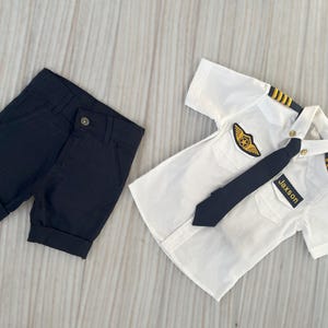 May include: A white pilot shirt with a black tie and epaulettes, paired with navy blue shorts. The shirt features a gold emblem and a name tag that reads "Jaxson".