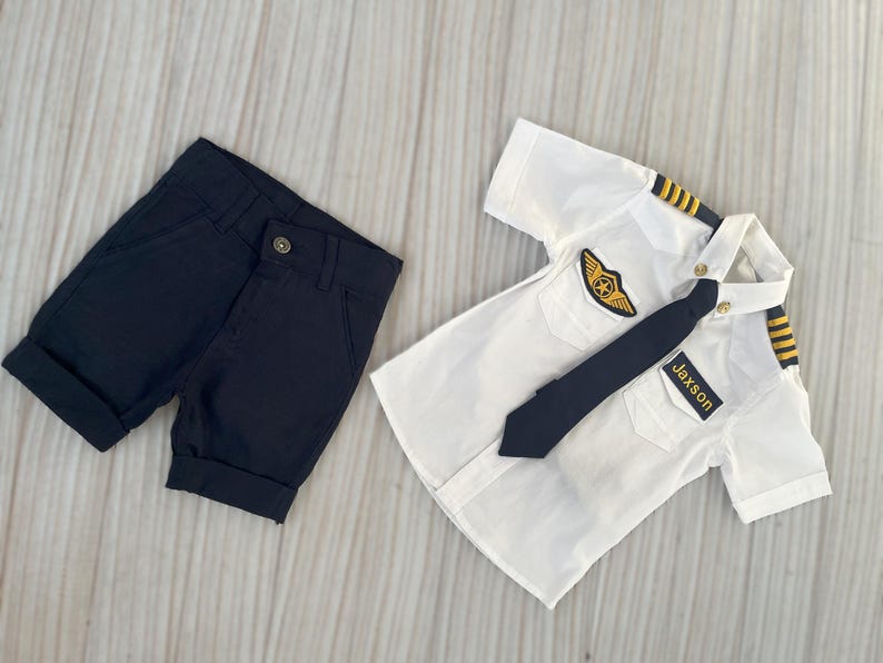 May include: A white pilot shirt with a black tie and epaulettes, paired with navy blue shorts. The shirt features a gold emblem and a name tag that reads "Jaxson".