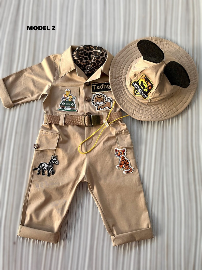 May include: A tan safari jumpsuit with a yellow belt and multiple animal patches. The jumpsuit has a name tag that says "Tadhg". The outfit is paired with a tan safari hat with black Mickey Mouse ears.