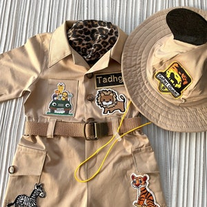 May include: A tan safari jumpsuit with a brown belt and several animal patches. The jumpsuit has a pocket on the left leg with a zebra patch. The right leg has a tiger patch. The jumpsuit also has a lion patch on the chest. The name "Tadhg" is written on a patch on the chest. A brown safari hat with a black brim and a patch that says "Safari Guide" is also included.