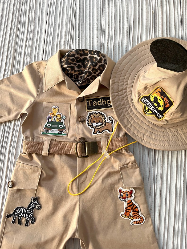May include: A tan safari jumpsuit with a brown belt and several animal patches. The jumpsuit has a pocket on the left leg with a zebra patch. The right leg has a tiger patch. The jumpsuit also has a lion patch on the chest. The name "Tadhg" is written on a patch on the chest. A brown safari hat with a black brim and a patch that says "Safari Guide" is also included.