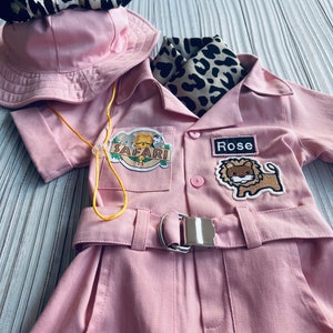 May include: Pink safari jumpsuit with a lion patch, a safari patch, and a name tag that says "Rose". The jumpsuit has a yellow lanyard, a brown and black leopard print scarf, and a pink hat with black mouse ears and a brown and black leopard print bow.