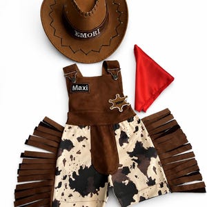 Op de afbeelding: Een cowboykostuumset met een bruine hoed met geborduurd "EMORI", een bruine en koeienprint overall met "Maxi" op de voorkant, een sheriff-badge en een rode bandana. De overall heeft bruine franjes.