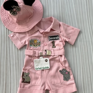 May include: Pink safari outfit for a baby or toddler, featuring a hat with Mickey Mouse ears, a safari shirt with a name tag that says "Savannah", and a safari guide badge. The outfit has several patches with animal designs, including a lion, an elephant, and a koala.