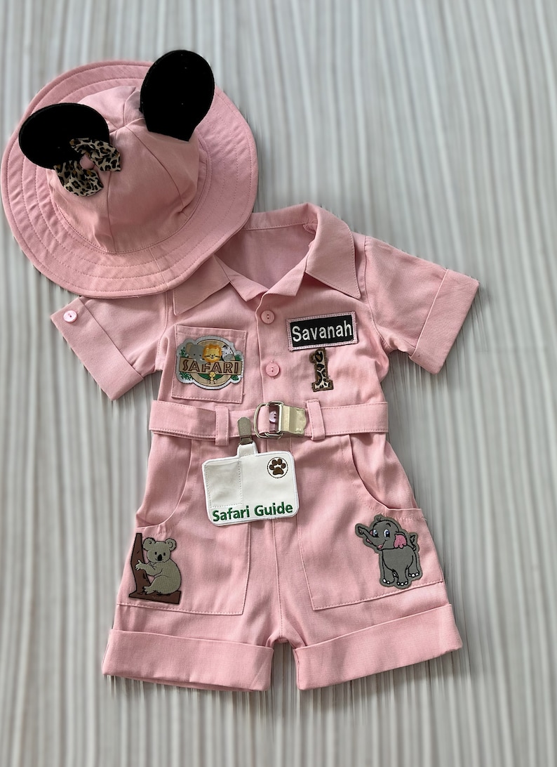 May include: Pink safari outfit for a baby or toddler, featuring a hat with Mickey Mouse ears, a safari shirt with a name tag that says "Savannah", and a safari guide badge. The outfit has several patches with animal designs, including a lion, an elephant, and a koala.