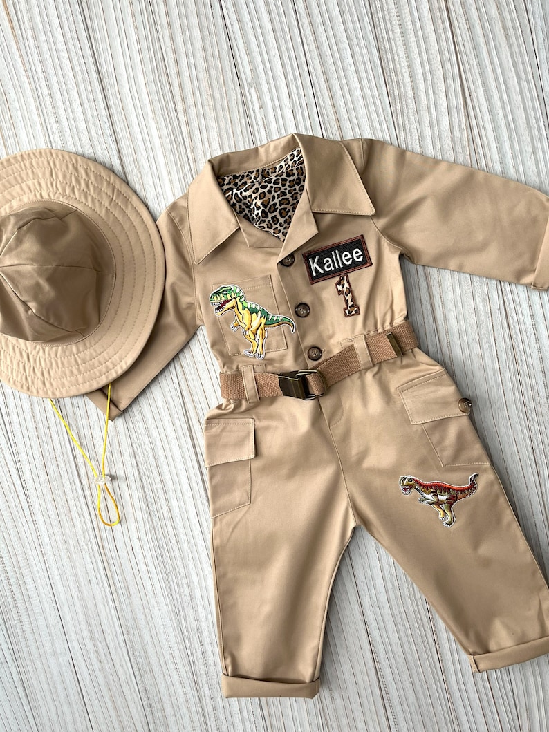 Pu&ograve; includere: Una tuta da safari beige con una cintura marrone, due toppe di dinosauro e un'etichetta con il nome "Kallie". La tuta &egrave; abbinata a un cappello da safari beige.