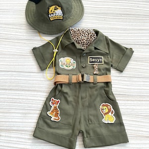 May include: A green safari jumpsuit with a brown belt, a tiger and lion patch, and a name tag that says "Sevyn". The jumpsuit has a safari hat with Mickey Mouse ears and a "Safari Tour Guide" patch.