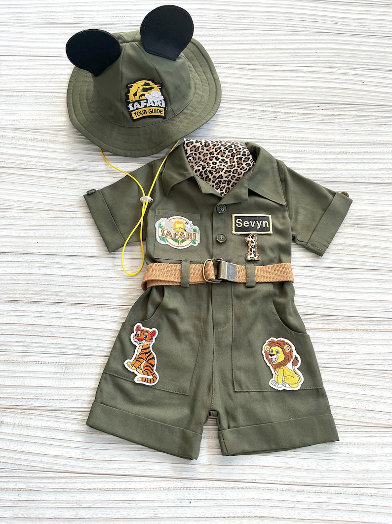 May include: A green safari jumpsuit with a brown belt, a tiger and lion patch, and a name tag that says "Sevyn". The jumpsuit has a safari hat with Mickey Mouse ears and a "Safari Tour Guide" patch.
