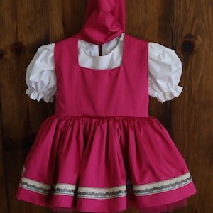 K&ouml;nnte beinhalten: Ein rosa Kleid mit einer wei&szlig;en Bluse und einem passenden rosa Hut. Das Kleid hat einen ger&uuml;schten Rock und einen wei&szlig;en Besatz um den Ausschnitt. Das Kleid ist mit einem gemusterten Band am Saum verziert.