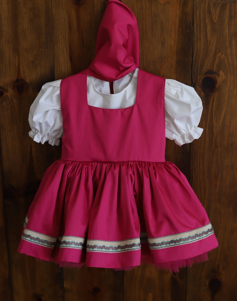 K&ouml;nnte beinhalten: Ein rosa Kleid mit einer wei&szlig;en Bluse und einem passenden rosa Hut. Das Kleid hat einen ger&uuml;schten Rock und einen wei&szlig;en Besatz um den Ausschnitt. Das Kleid ist mit einem gemusterten Band am Saum verziert.