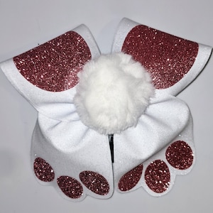 May include: White glitter bunny ears headband with a pink glitter pom pom in the center. The ears have pink glitter paw prints on the tips.