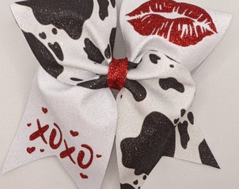 Peut inclure: Un nœud de cheerleading en tissu imprimé vache noir et blanc avec un centre en paillettes rouges et des lèvres rouges en paillettes d'un côté et le mot "XOXO" en paillettes rouges de l'autre côté.