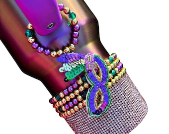 May include: A purple and gold tumbler with a beaded strap and a sequined mask charm. The tumbler is covered in rhinestones.