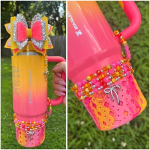 May include: A Stanley tumbler with a pink and yellow ombre design. The tumbler is decorated with a large glitter bow, beaded accents, and a decorative bow charm. The handle and base are also pink.