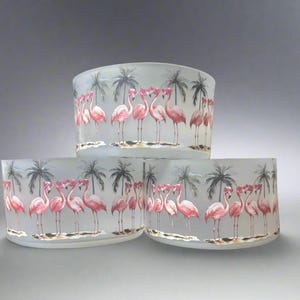 May include: Three frosted glass candle holders with a tropical flamingo design. Each holder features pink flamingos, palm trees, and a sandy beach scene. The candle holders are stacked, showcasing the design.