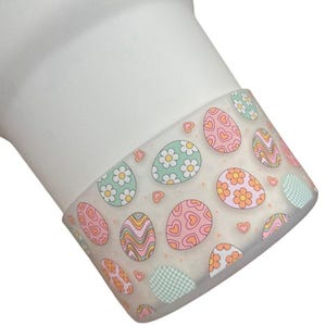 May include: White ceramic mug with a clear plastic band decorated with a pattern of pastel pink, green, and orange Easter eggs. The eggs have different designs, including hearts, flowers, and swirls.