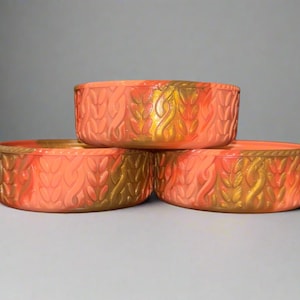 May include: Three coral-colored, round bowls with a textured, woven design and gold accents. The bowls are stacked, with the top bowl slightly overlapping the other two. The bowls have a decorative, artistic appearance.