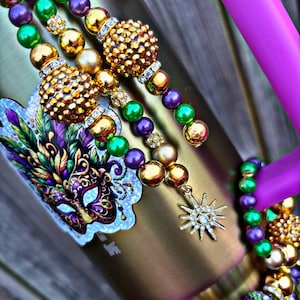 May include: A gold, purple, and green beaded necklace with a star charm and a Mardi Gras mask sticker on a metallic tumbler.