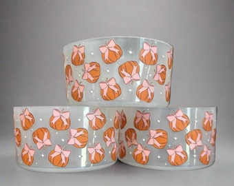 May include: Three clear glass bowls stacked, each adorned with a repeating pattern of orange pumpkins tied with pink bows and small white star accents. The bowls have a rounded shape and a frosted appearance.