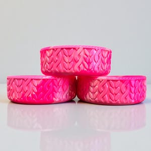 May include: Three bright pink, round containers stacked together. Each container has a textured, woven pattern around the sides. The containers are sitting on a reflective surface.