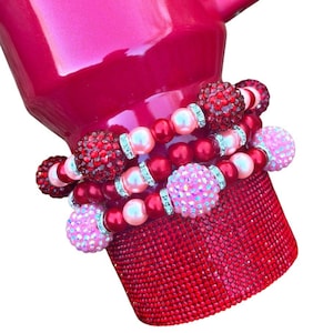 May include: A red and pink beaded bracelet set with rhinestone accents. The bracelets are wrapped around a red, sparkly mug.