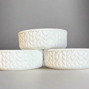 May include: Three white ceramic bowls with a textured leaf pattern. The bowls are stacked, with two on the bottom and one on top. The bowls are on a white surface with a gray background.