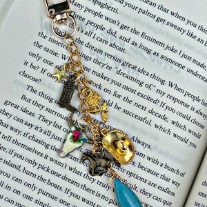 May include: A gold-toned metal keychain featuring a lobster clasp and a chain adorned with multiple charms including a small star, a cowboy boot, a rose, a steer skull with floral details, a cowboy hat, a horse, and a teardrop-shaped turquoise stone.