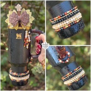 May include: Black insulated tumbler with a snakeskin texture, adorned with a gold and glitter bow, and beaded accents. The tumbler features a handle and is embellished with multiple beaded bracelets. The Stanley logo is visible.