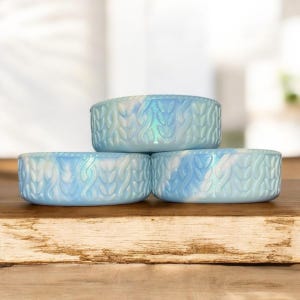 May include: Three light blue ceramic bowls stacked on a wooden surface. Each bowl has a textured, woven-like pattern and a marbled effect with white and iridescent accents. The bowls are round and shallow.