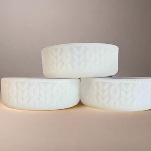 May include: Three off-white, round containers stacked. Each container features a raised, textured pattern resembling a cable knit design. The containers are evenly lit, with soft shadows, and are set against a neutral background.