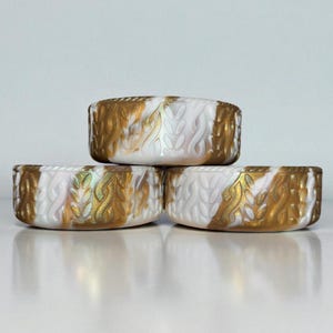 May include: Three ceramic bowls with a white and gold design. The bowls have a textured pattern with gold accents and a white background. The bowls are stacked on top of each other.