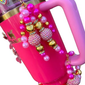 May include: A pink stainless steel tumbler with a pink handle and a gold lid. The tumbler is decorated with pink, gold, and clear beads. The beads are arranged in a cascading pattern. The tumbler is a popular item for everyday use.