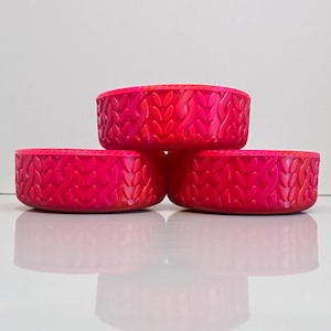 May include: Three stacked, round, hot pink bowls with a textured, woven pattern. The bowls are arranged in a pyramid shape, with the bottom two bowls supporting the top bowl. The bowls are made of a solid material and have a smooth, reflective surface.