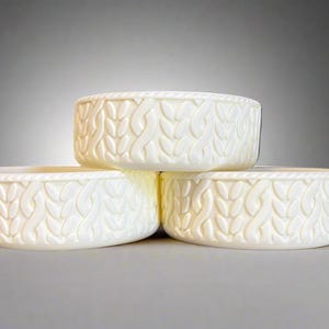 May include: Three off-white, round bowls stacked together. Each bowl has a textured leaf pattern around the sides and a rope-like design at the top. The bowls are made of a smooth material.